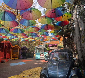 A charming and whimsical outdoor space adorned with a canopy of colorful suspended umbrellas, featuring a classic blue Volkswagen Beetle on a brick-paved path with a yellow brick road design. The scene is set in a quaint area with a cozy corner food stand, creating a magical and picturesque atmosphere that invites visitors to enjoy and explore. balat town