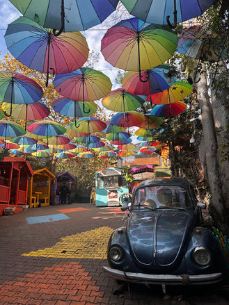 A charming and whimsical outdoor space adorned with a canopy of colorful suspended umbrellas, featuring a classic blue Volkswagen Beetle on a brick-paved path with a yellow brick road design. The scene is set in a quaint area with a cozy corner food stand, creating a magical and picturesque atmosphere that invites visitors to enjoy and explore. balat town