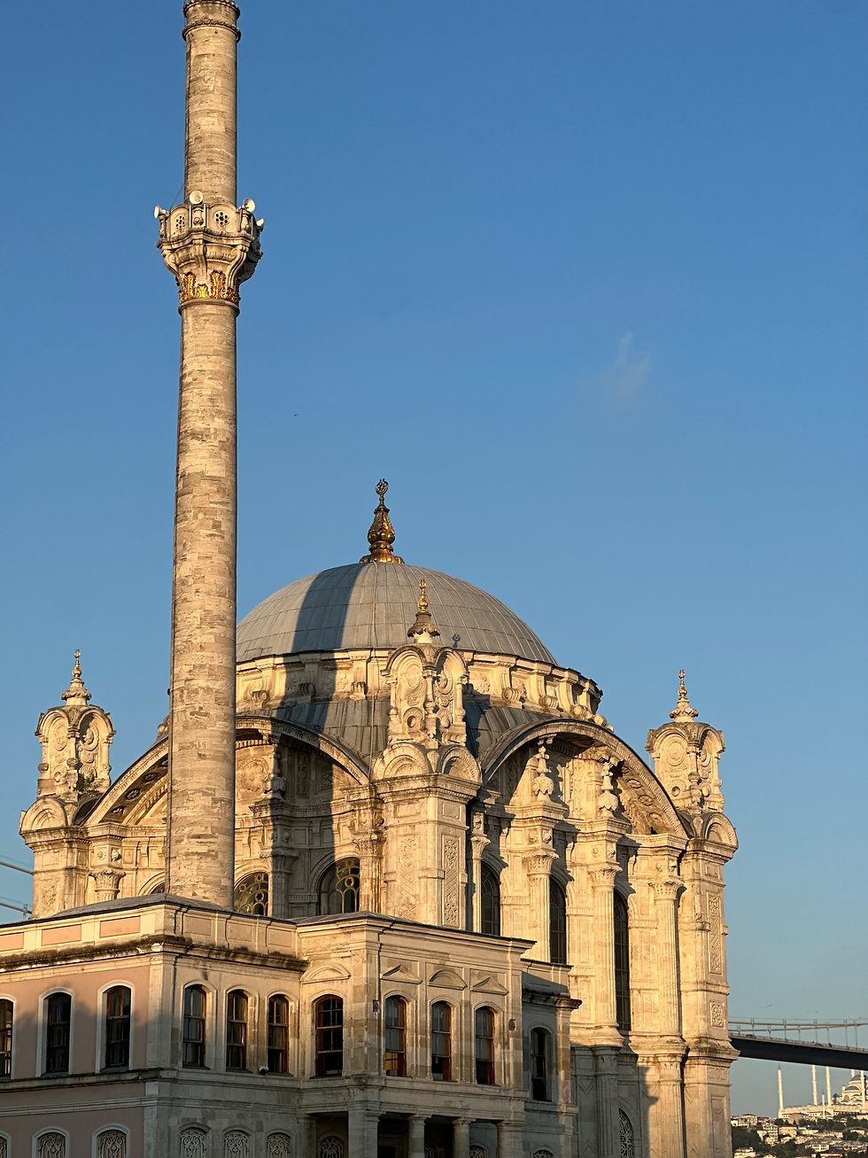 Ornate mosque with a large dome and minaret under a clear blue sky. Sunlit facade with intricate details. Bridge visible in the distance.