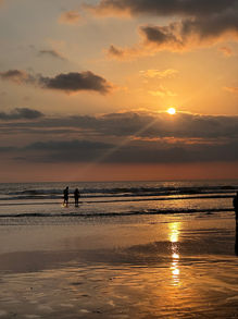 A breathtaking sunset on the beach with the sun dipping below the horizon, casting a warm golden glow over the sky and the sea, as silhouettes of people enjoy the peaceful evening by the water's edge.