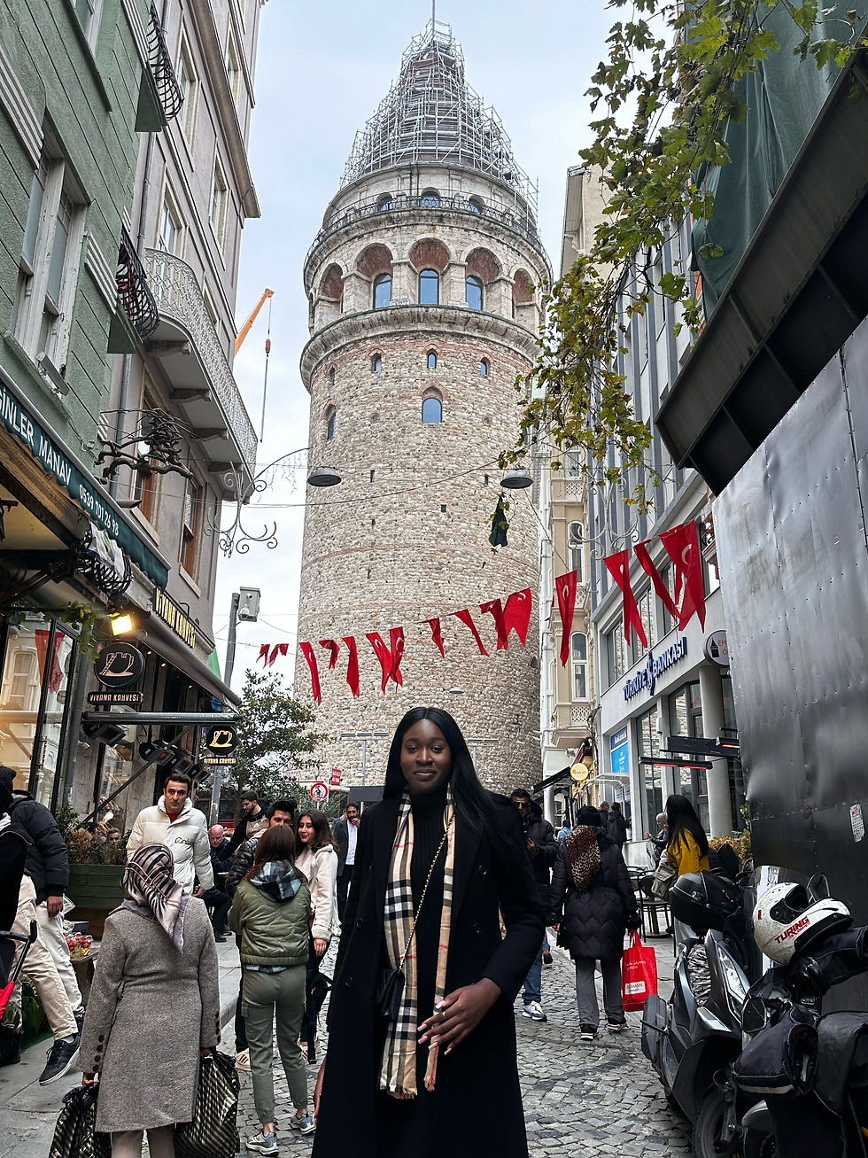 Woman in black coat stands on cobbled street with crowd near Galata Tower, Istanbul. Red flags hang above; a lively urban scene.