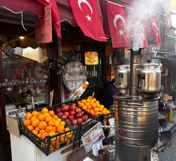 A vibrant street food stall in Turkey with Turkish flags, a traditional tea-making machine releasing steam, a display of fresh oranges and pomegranates, and signs offering local delicacies like baklava and pomegranate juice. The setting has an authentic Turkish ambiance with a glimpse of daily life and culture.
