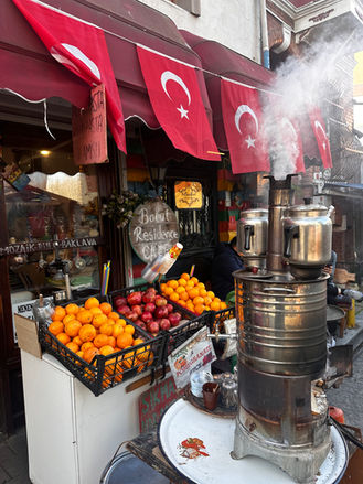 A vibrant street food stall in Turkey with Turkish flags, a traditional tea-making machine releasing steam, a display of fresh oranges and pomegranates, and signs offering local delicacies like baklava and pomegranate juice. The setting has an authentic Turkish ambiance with a glimpse of daily life and culture.