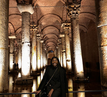 A joyful visitor smiles in front of the historic Basilica Cistern in Istanbul, with its ancient, dimly lit stone columns and arches reflecting in the still water, giving a glimpse into the grandeur of Byzantine architecture.