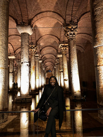 A joyful visitor smiles in front of the historic Basilica Cistern in Istanbul, with its ancient, dimly lit stone columns and arches reflecting in the still water, giving a glimpse into the grandeur of Byzantine architecture.