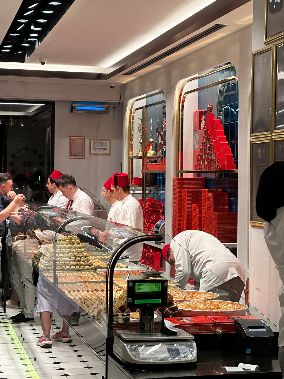 Bakers in red hats serve pastries in a well-lit bakery with red accents. Pastries are displayed under glass. Customers observe intently.
