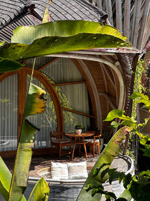 A cozy and inviting tropical retreat corner framed by lush greenery and banana leaves, with a circular bamboo structure housing comfortable seating and a wooden table, embodying a peaceful and natural environment for relaxation and conversation.