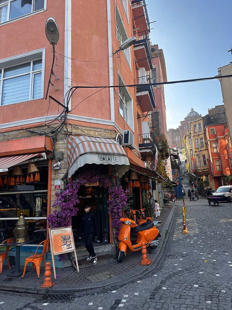 Street scene with an orange building and a cafe entrance adorned with purple flowers. An orange scooter is parked outside. A person is walking by.