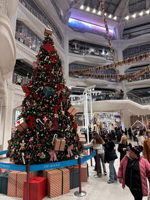 A festive holiday scene inside a bustling shopping center adorned with a towering Christmas tree, beautifully decorated with red and gold ornaments, ribbons, and wrapped gifts at its base. Shoppers enjoy the seasonal decor, which adds to the cheerful atmosphere of the multi-level retail space during the Christmas shopping season.