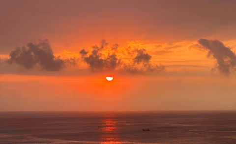 A stunning ocean sunset view with the sun partially hidden by clouds, casting vibrant hues of orange and red across the sky and reflecting off the calm sea, creating a tranquil and picturesque moment at dusk.