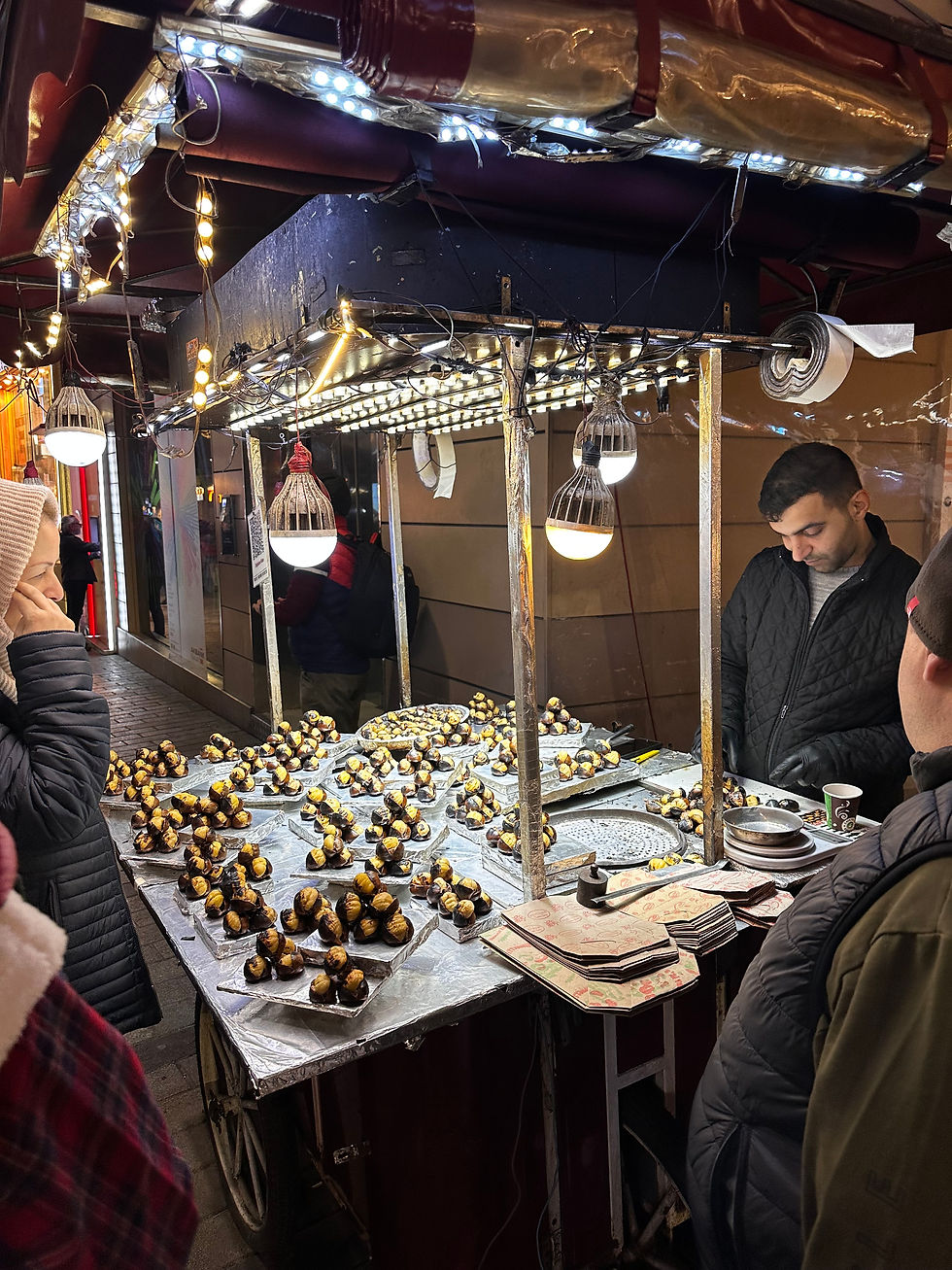 Street vendor selling roasted chestnuts under illuminated stall with string lights. People in jackets waiting. Cozy evening atmosphere.