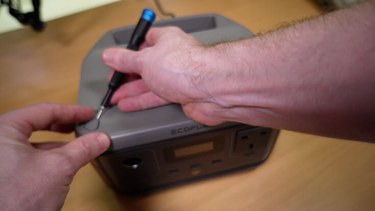 Hands using a screwdriver on a grey device labeled "ECOFLOW" on a wooden table background. The setting appears technical and focused.