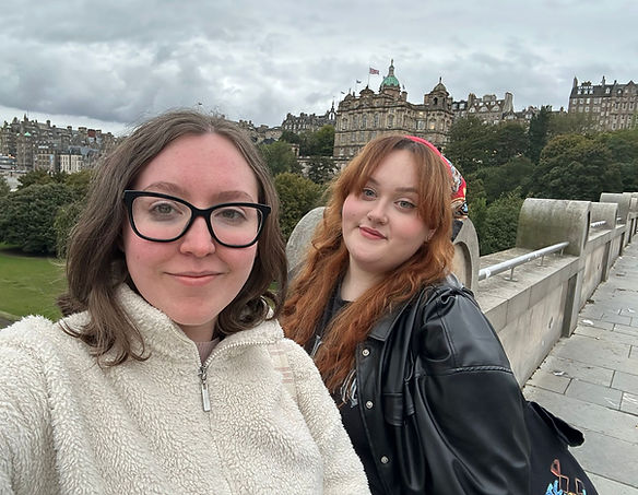 Two young women stand in front of a castle. The left one is a brunette, the right one has ginger hair.