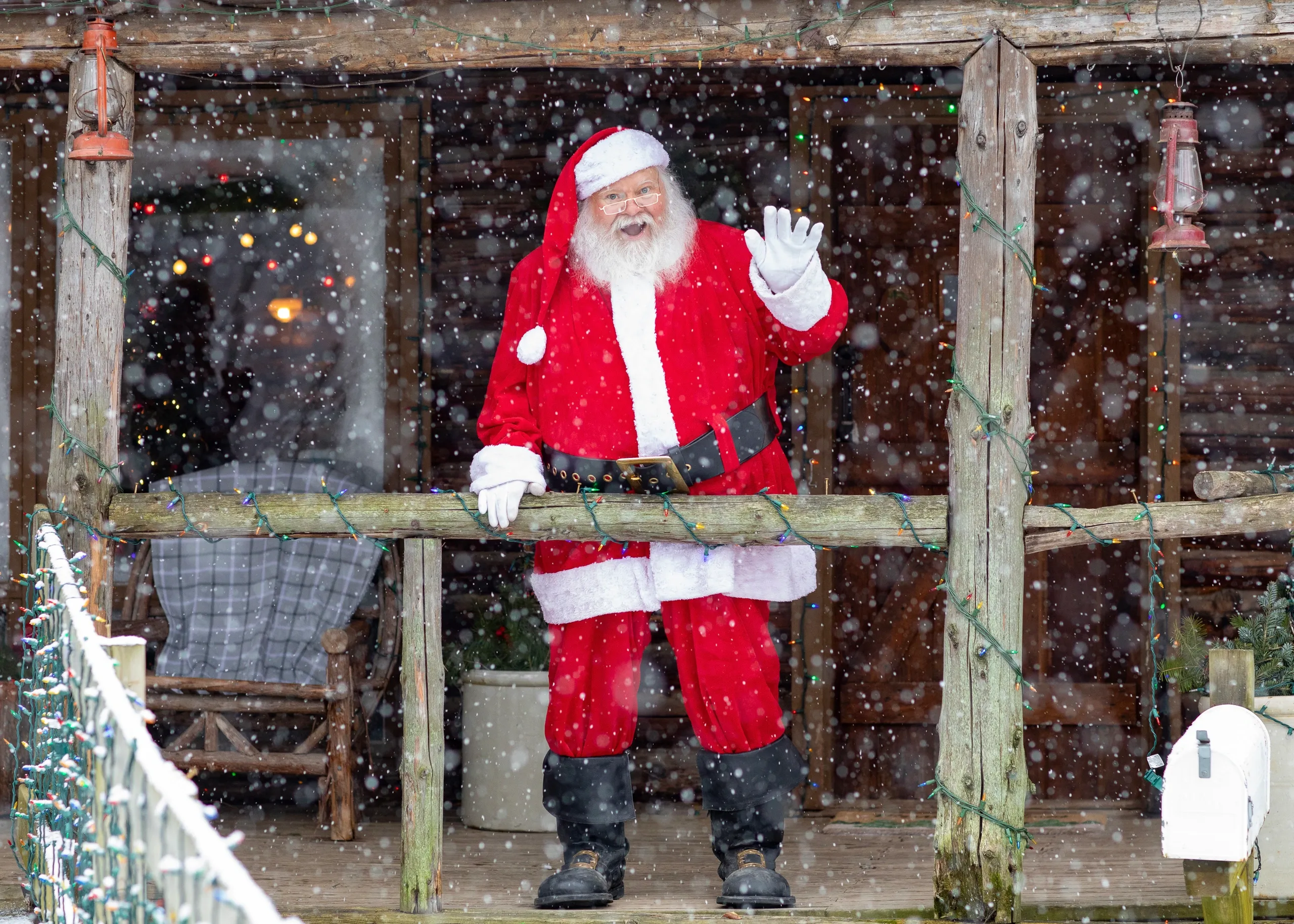 Santa Claus waves from the front porch of a snow-covered log cabin, surrounded by festive decorations during the Santa Experience at Peacock Road Family Farm.