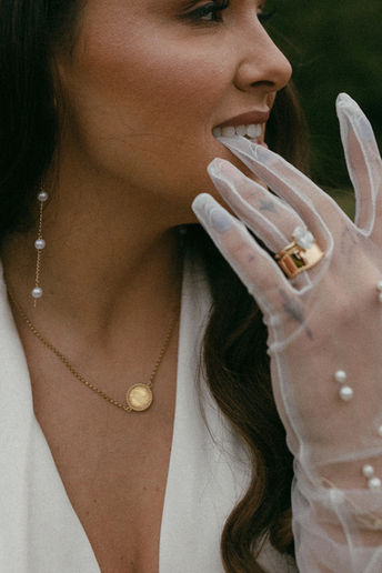 Woman wearing white gown and sheer, white gloves with a gold necklace taking off one of the gloves with her teeth