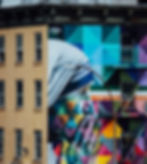 A striking street art mural depicting a person's face in profile with their hand gently touching their face, painted in soft grays against a backdrop of vibrant geometric shapes in blues, purples, and yellows. The contrast between the monochromatic figure and colorful background creates a powerful metaphor for the interplay between inner reflection and the diverse experiences of neurodivergent minds. The mural spans multiple stories of an urban building with fire escapes.