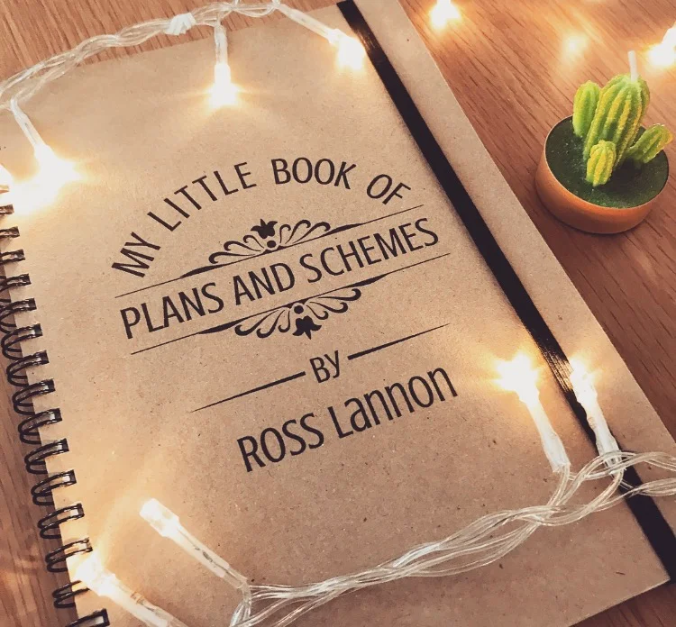 A notebook with the frontpage titled "My little book of plans and schemes, by Ross Lannon" positioned next to a cacti candle and fairy lights