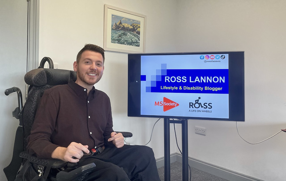 Ross sat beside a TV screen displaying a powerpoint presentation with his name and blog logo on