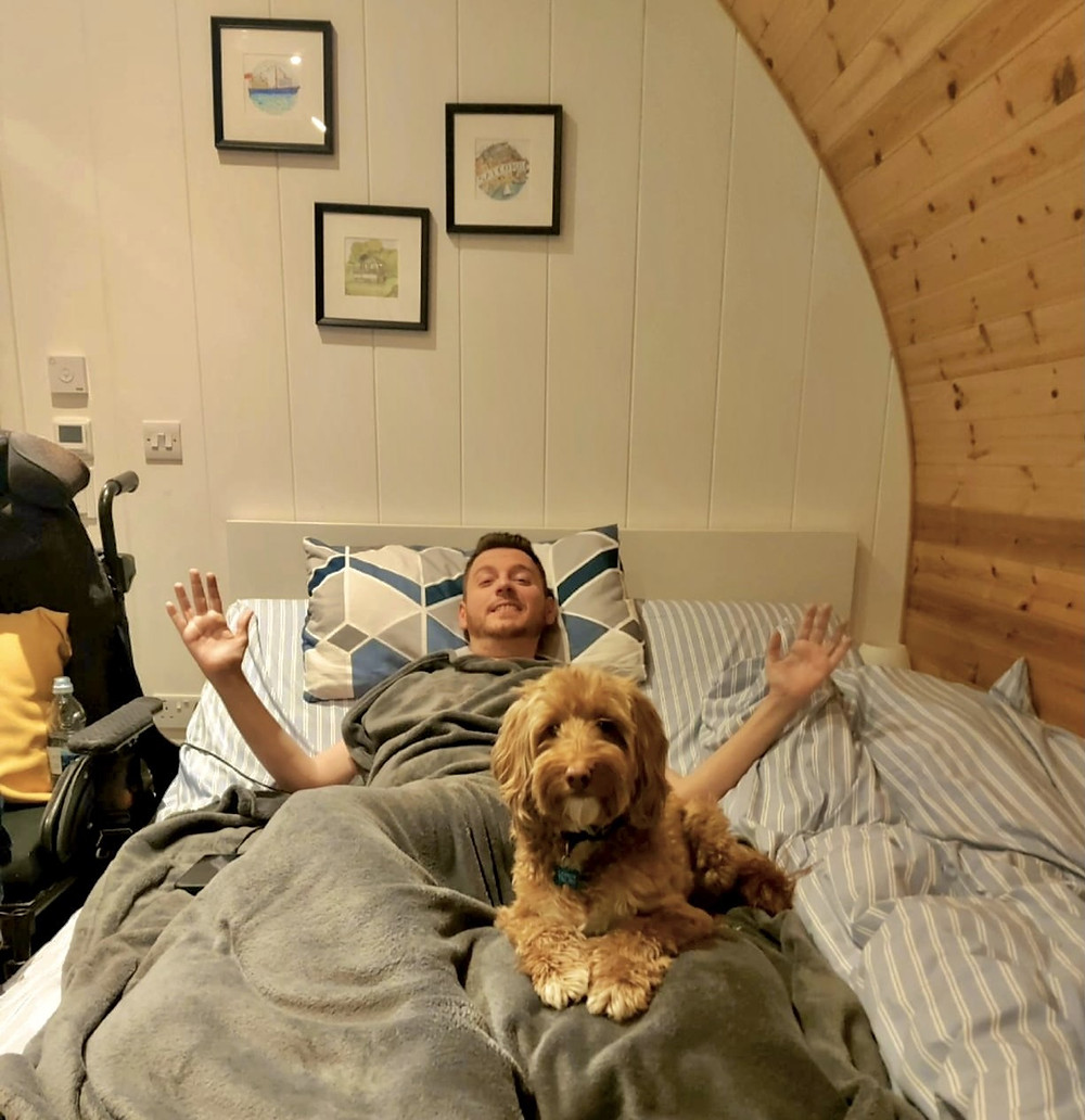 Ross laying in the bed under a blanket, with his dog Ralph sat on his lap