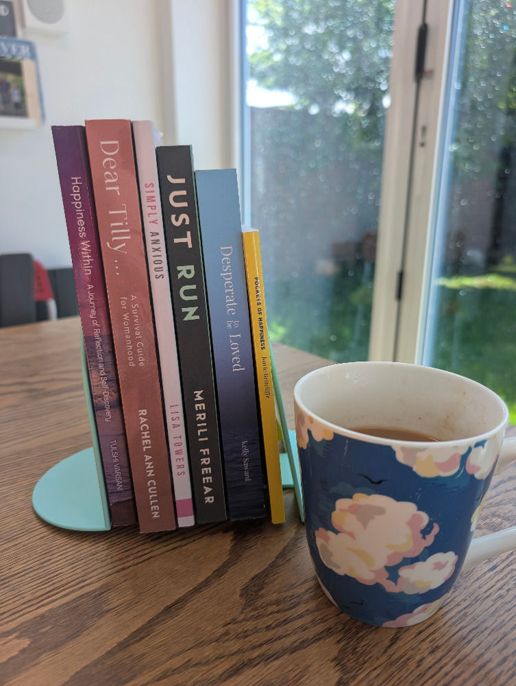 Books on a wooden table with a blue, cloud-patterned mug beside them. Sunlight streams through a window, creating a calm atmosphere.