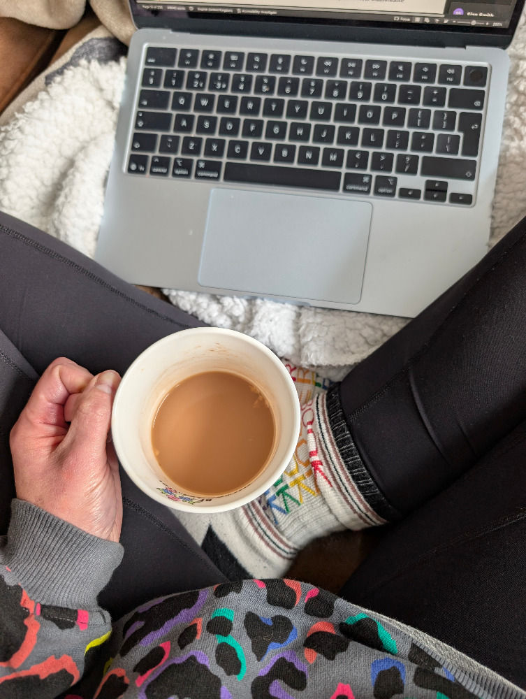 A person sits with a laptop on their lap, holding a cup of coffee. They wear colorful socks and a vibrant sweater with a rainbow leopard print, creating a cozy vibe.