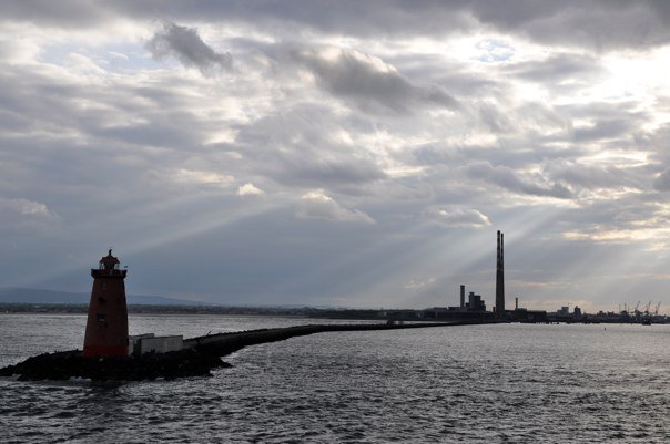 Dublin Bay lighthouse on a dramatic overcast day, with rays of sunlight breaking through the clouds above the water and industrial chimneys in the distance.