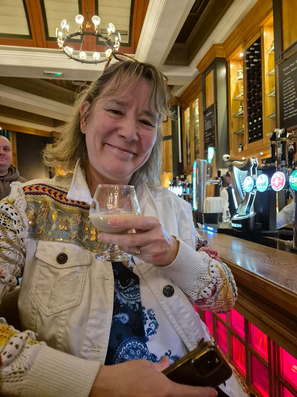 Tanya smiling and raising a glass of Baileys at a pub bar, wearing a white embroidered jacket, looking relaxed and joyful.