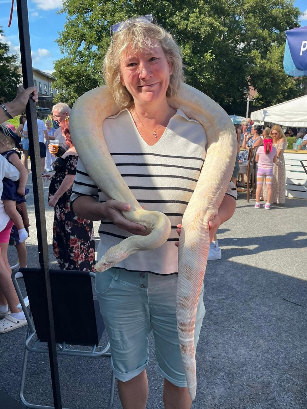 Woman smiles holding a large albino snake at an outdoor event. People in background. Blue sky and trees set the scene. Casual atmosphere.
