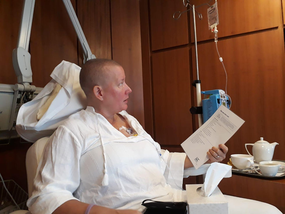 Tanya during chemotherapy treatment, seated in a hospital recliner with an IV drip, wearing a white robe, head shaved, calmly reading a menu, with a tea service beside her.
