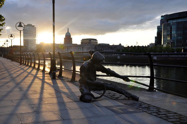 A bronze sculpture of a figure pulling a rope on the banks of the River Liffey in Dublin at sunrise, with golden light flooding the city skyline behind.