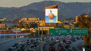 Traffic flows on a highway with a mountain backdrop. A billboard shows a baseball and adverts for LA Dodgers and a stadium.