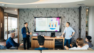 Men present to seated group in modern office. Graph on screen shows YTD growth. Room has floral wallpaper, relaxed atmosphere.