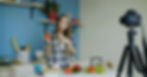 Woman in plaid shirt holding a bell pepper, smiling in a kitchen studio. Camera on tripod, blue panel wall, cheerful mood.