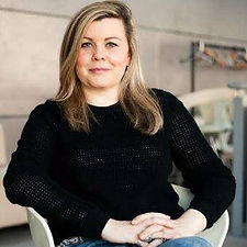 Woman with mid length dark blond hair sits in a chair looking directly at the camera, she's smiling