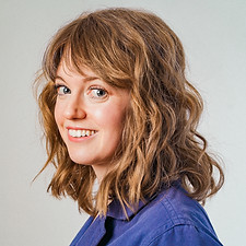 Megan is smiling at the camera with her body facing toward the left. She is a white woman with wavy, blonde, shoulder-length hair, and she is wearing a purple jumpsuit.