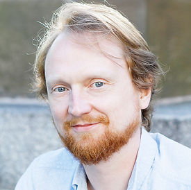 A man with a reddish beard and hair looks towards the camera. He is wearing a light blue shirt.