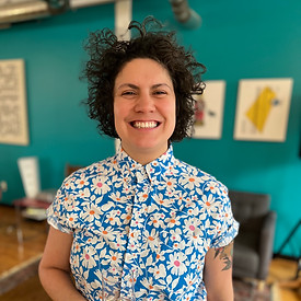 The image shows a person standing indoors, smiling broadly at the camera. They have short, curly dark hair and are wearing a vibrant, short-sleeved button-up shirt with a bold floral pattern�—white daisies with orange centers on a blue background. The setting appears to be a cozy, modern space with teal-colored walls, framed artwork, and casual furniture including a chair, table, and a patterned rug. The atmosphere feels cheerful and relaxed.