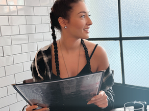 Woman with long braided hair sitting by a window inside The Smith restaurant in New York City, holding a menu in soft natural light with white tiled walls behind her.