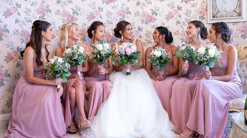 Bride in white and six bridesmaids in pink dresses sit smiling, holding bouquets. Floral wallpaper and vintage decor in the background.