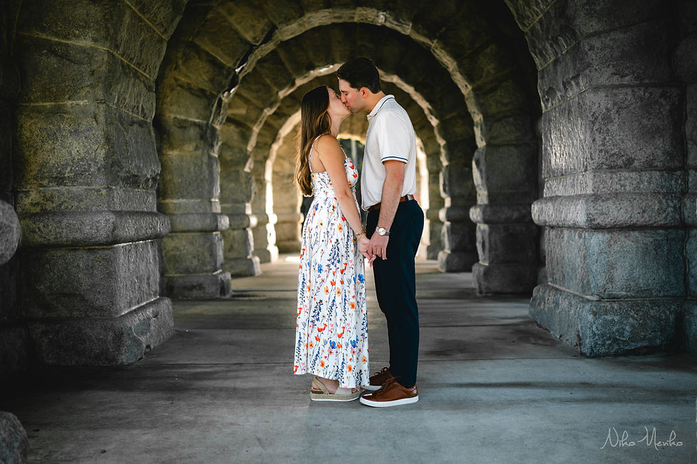 Couple kissing under a stone archway, hand in hand; she wears a floral dress, he wears a white polo, in soft warm light.