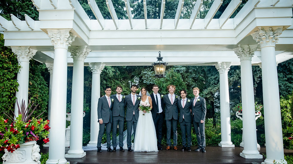 Bride with seven groomsmen in suits under a white pergola with columns. Lush greenery in the background sets a joyful wedding scene.
