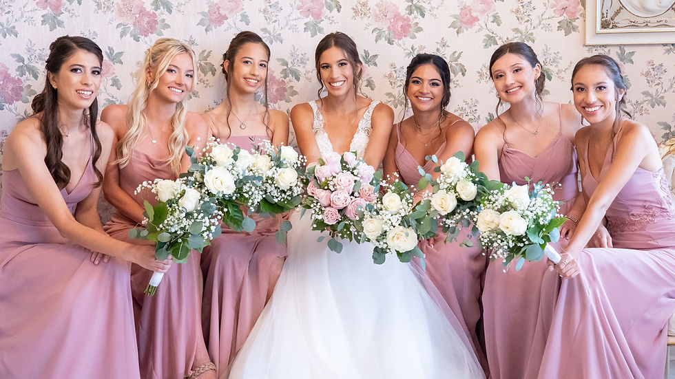Bride in white, surrounded by six bridesmaids in pink dresses, holding bouquets. Floral wallpaper background, joyful and smiling.
