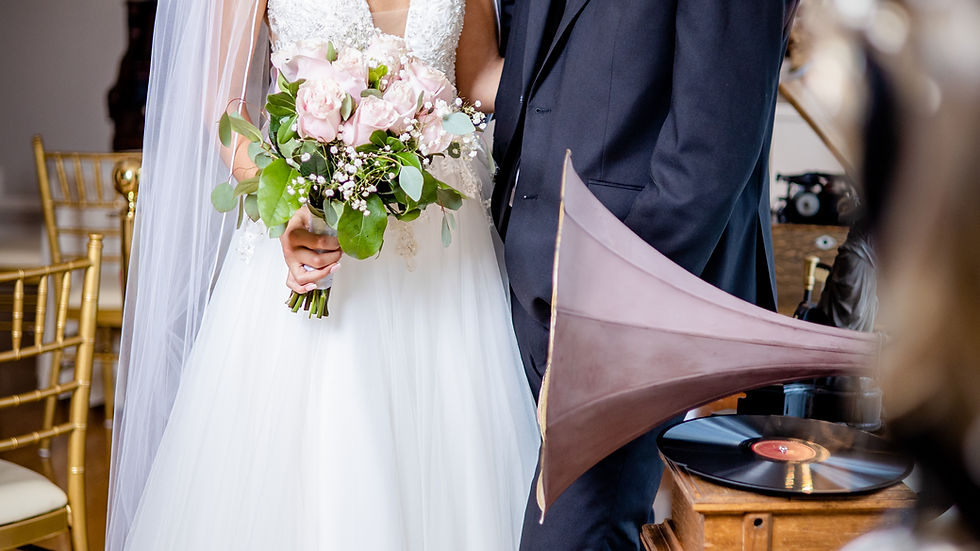 Bride in white dress and groom in suit smiling, holding pink bouquet. Elegant room with chandelier, vintage record player on side. Romantic mood.