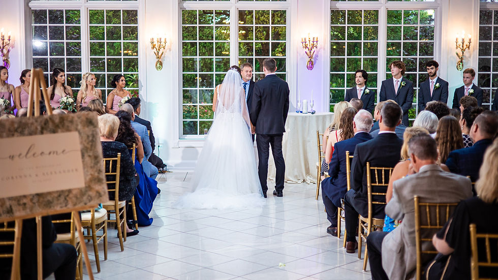 Bride and groom stand before officiant, holding hands in a bright hall with large windows. Guests seated on gold chairs, flowers visible.