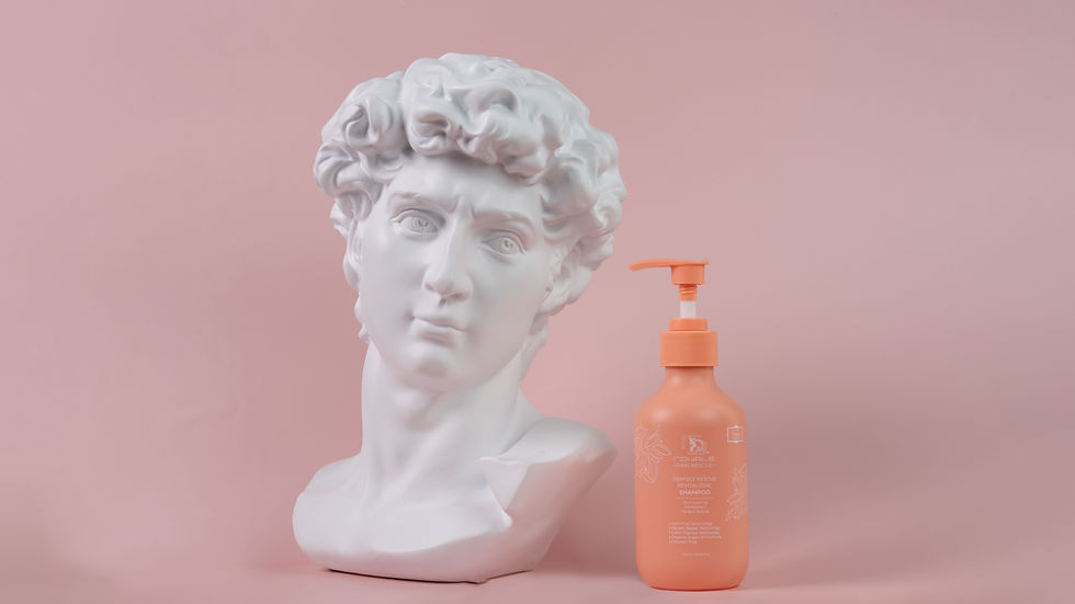 White marble bust beside an orange shampoo bottle on a pink background. The shampoo label features botanical designs and text.