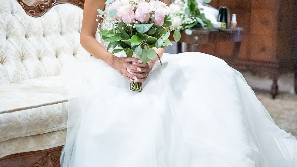 A bride in a white dress sits on a vintage sofa holding pink roses. A lamp and wooden dresser are in the background, creating an elegant mood.