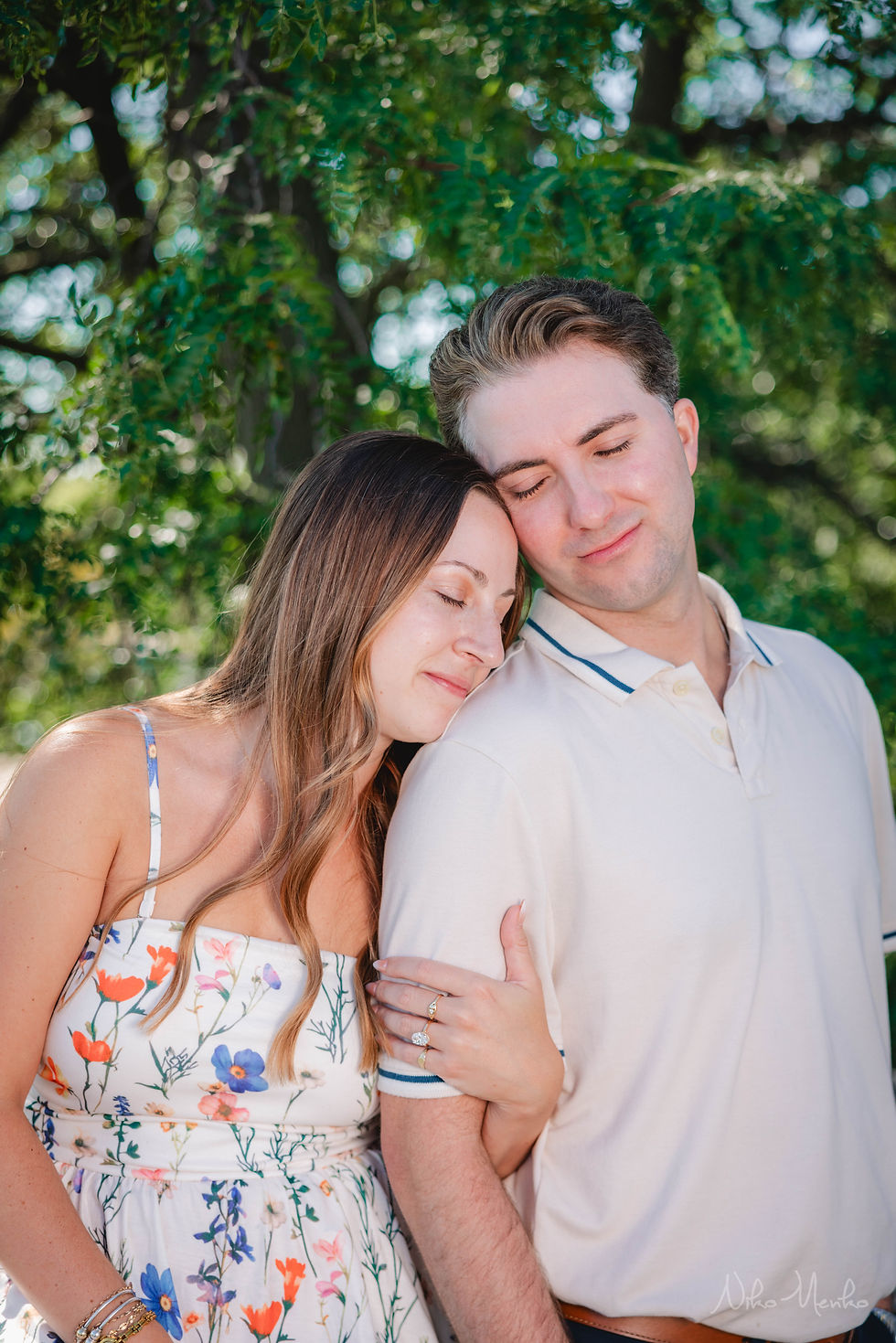 Smiling couple with eyes closed, embracing in front of leafy green trees; woman in floral dress, man in white polo.