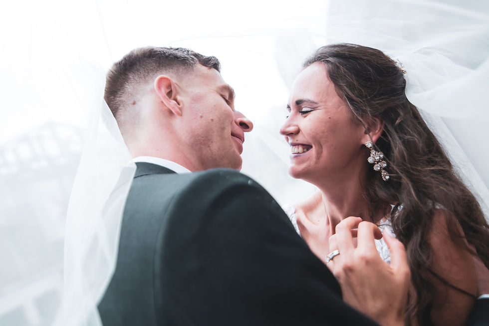 Bride and groom smiling joyfully under a veil, close-up shot. Bride wears sparkling earrings; soft, blurred background indicates wedding setting.