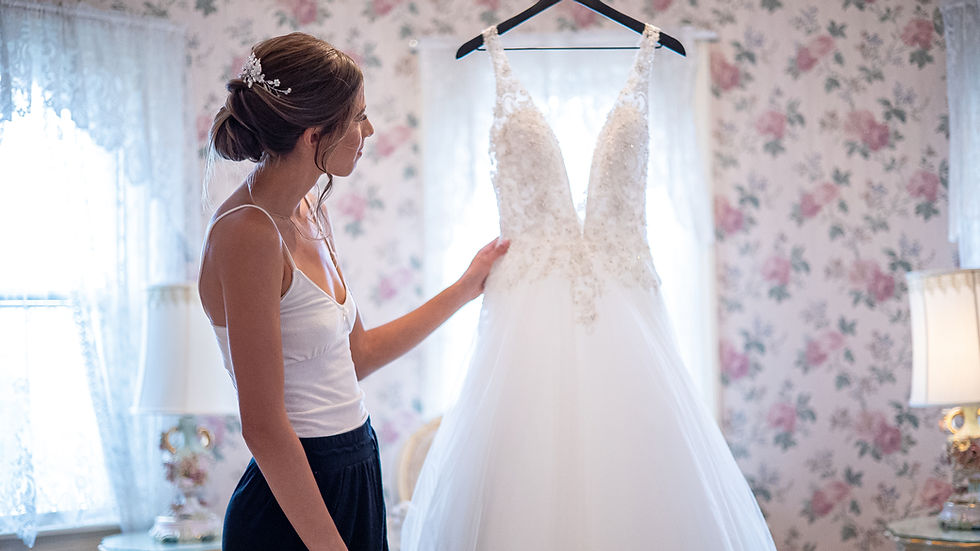 Woman in a tank top admiring a wedding dress on a hanger in a floral-patterned room with lace curtains, evoking a serene mood.