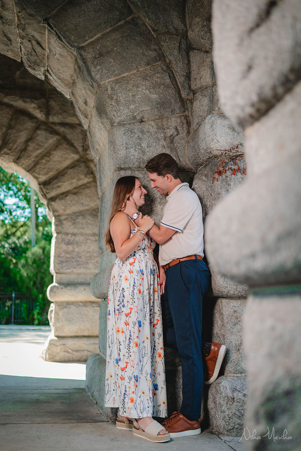 Smiling couple embrace under a stone archway, gazing at each other in a romantic outdoor setting.